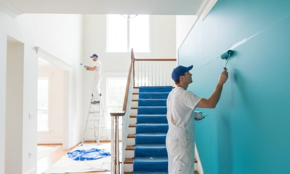 Two professional painters in white uniforms and blue caps painting a large, high-ceilinged interior entryway in a Connecticut home. One painter is on a ladder in the background, while the other applies teal paint to a long wall with a roller in the foreground. Blue protective film covers the stairs, and white drop cloths protect the hardwood floors.